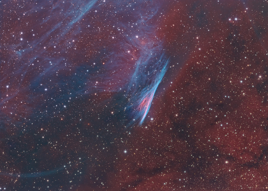 Vor einem Hintergrund aus dunkelbraunen Nebeln und vielen matten, gleichmäßig verteilten Sternen leuchtet ein blauer Nebel, der aussieht wie ein Streifen, der sich nach unten auffächert.