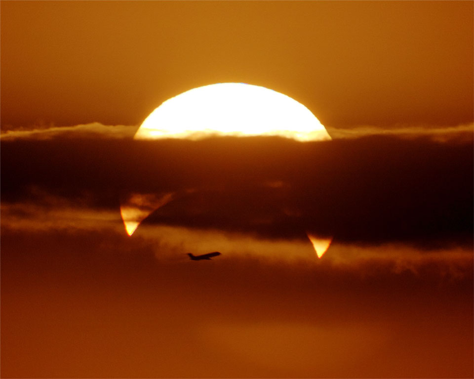 Hinter einer dicken Wolkenbank leuchtet die Sonne, teilweise vom Mond verdeckt, sie hat eine Sichelform. Darunter fliegt ein Flugzeug am orange gefärbten Himmel.