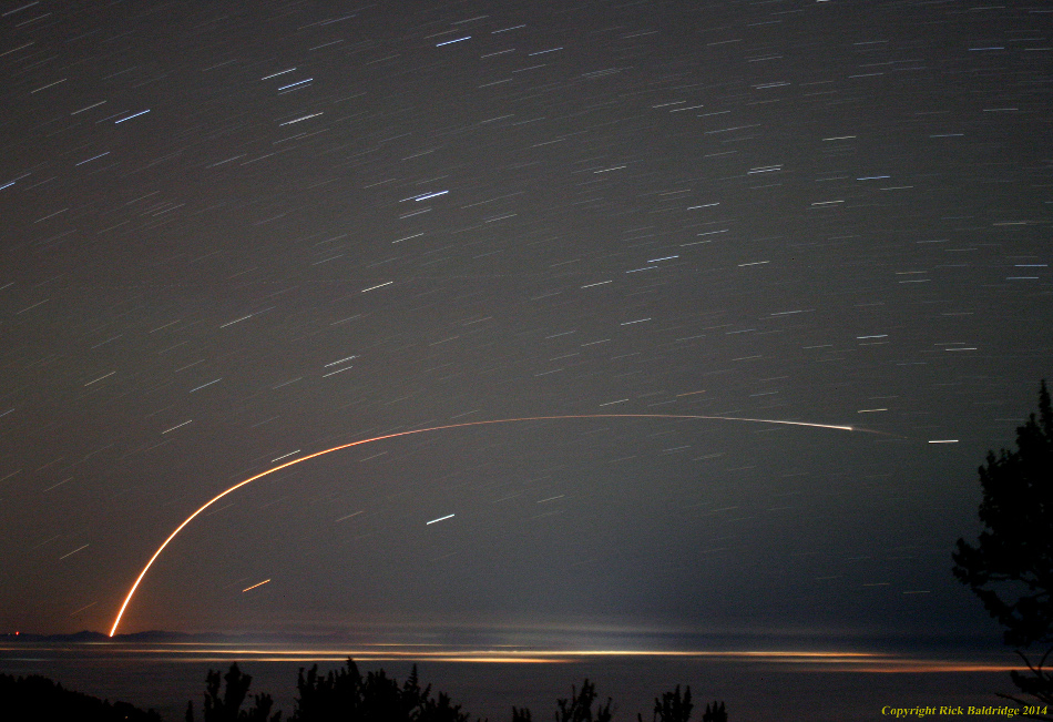 Eine startende Rakete malt einen hellen Bogen an den Nachthimmel. Im Hintergrund ziehen die Sterne Strichspuren am Himmel.