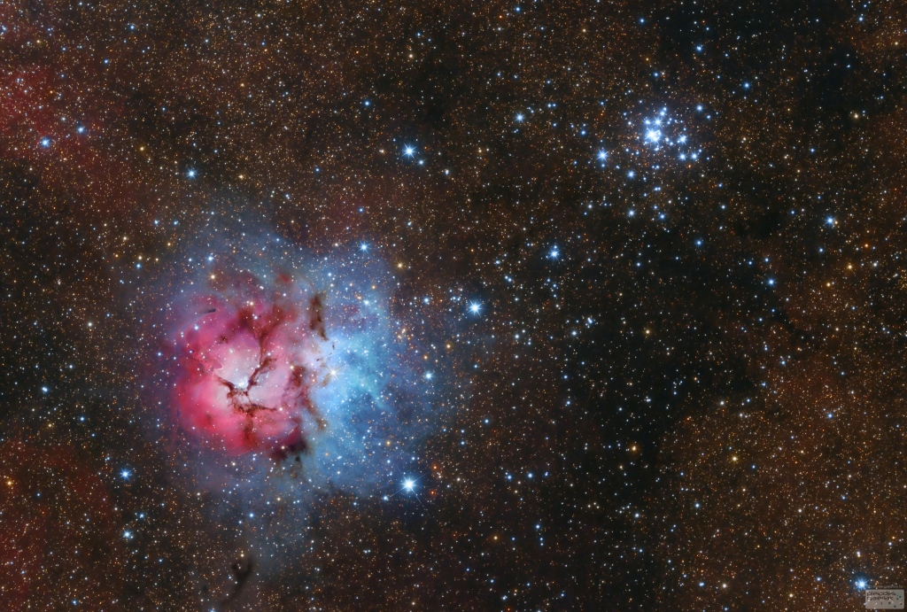 Links im Bild leuchtet der berühmte Trifidnebel, ein rosaroter Nebel, der von Staubwolken dreigeteilt ist, umgeben von einem blauen Nebel. Rechts darüber strahlt ein Sternhaufen. Im Hintergrund sind Sterne und kaum sichtbare Nebel verteilt.