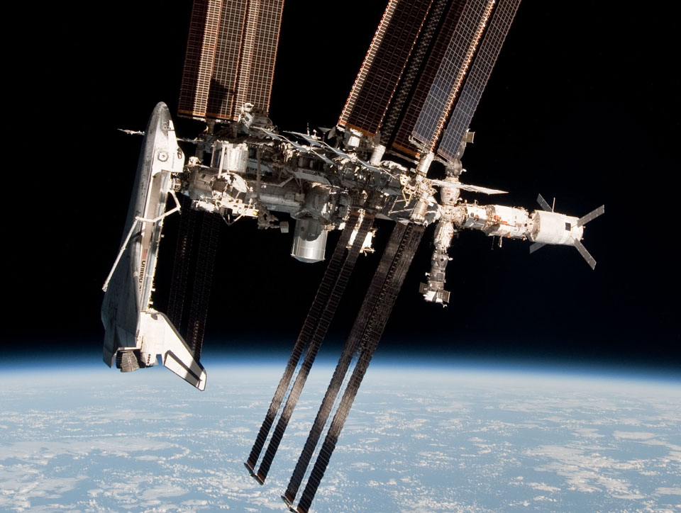 Über der Erde schwebt die  Internationale Raumstation mit Solarpaneelen und Modulen. Links ist eine Raumfähre angedockt, die Nase schaut nach oben. Unten wölbt sich die Erde mit blauen Ozeanen und weißen Wolken.