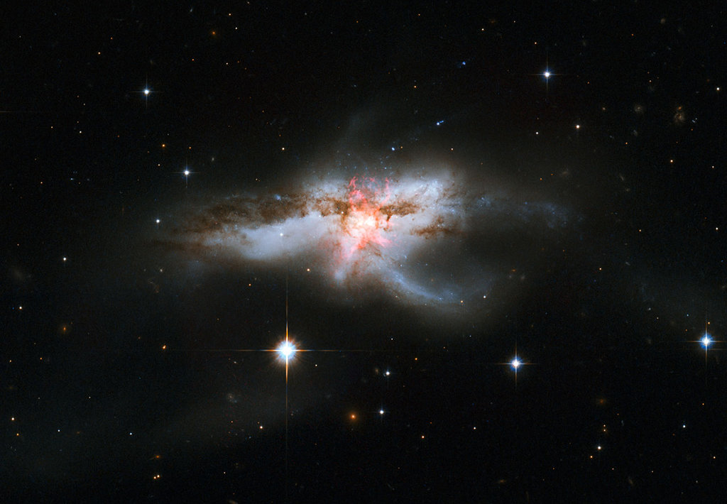 Mitten im Bild leuchtet eine Galaxie, deren Aussehen an eine Explosion erinnert. Im Inneren sind rote und dunkle Wolken über einem hellgelben Zentrum. Im Hintergrund leuchten nur wenige Sterne.