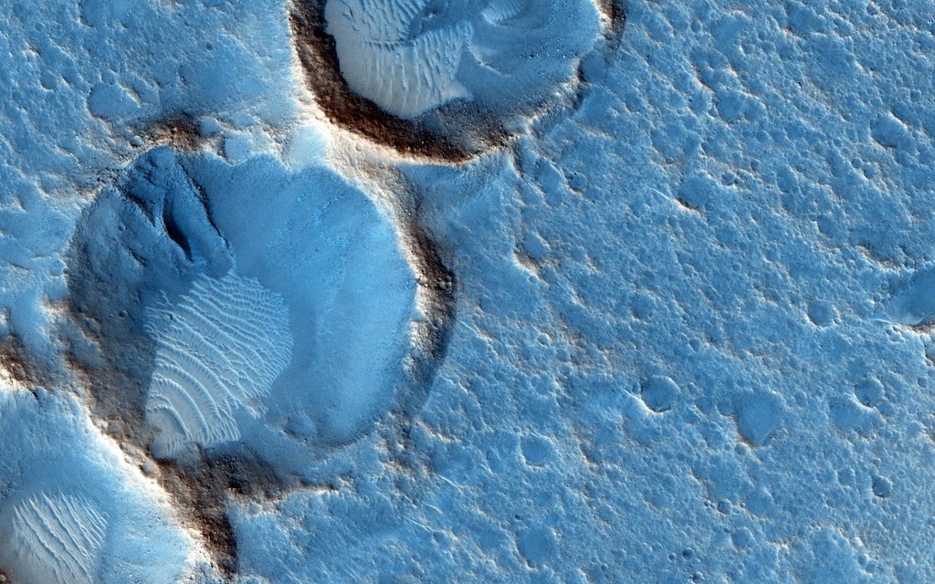 Auf einer blauen Planetenoberfläche mit vielen Kratern, die an zerfallenen Badeschaum erinnern, haben sich drei größere, tiefere Krater mit hohen Wällen eingegraben. Das Blau sind Falschfarben.