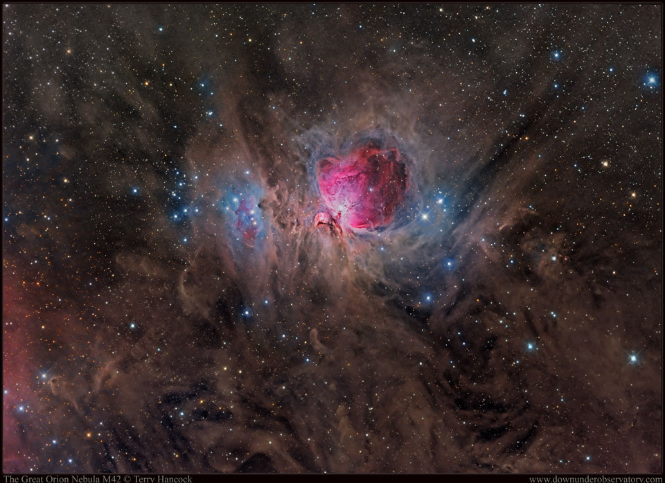 Umgeben von bräunlichen Staubwolken leuchtet in der Bildmitte der magentafarbene Orionnebel in einer Staubhöhle. Links davon ist ein Sternhaufen, umgeben von blauen Reflexionsnebeln.