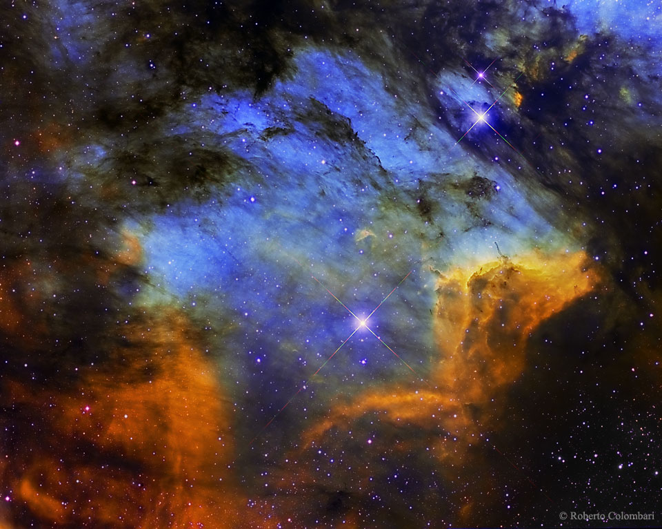 Das farbige Bild leuchtet in der Mitte blau, umgeben von dunklen Nebeln, unten sind einige orange leuchtende Wolken zu sehen. Das ganze Bild ist von markanten dunklen Staubwolken durchzogen.
