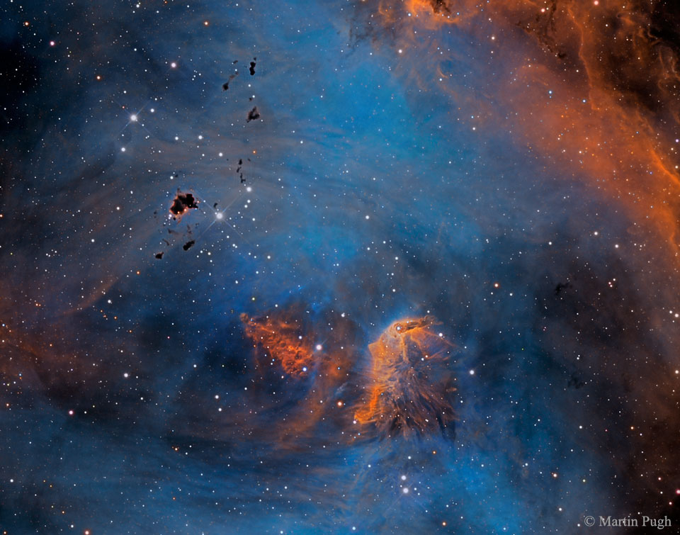 Vor einem blauen Hintergrund schweben einige orange Nebel und dunkle Wolkenfetzen aus dichtem Staub, links unten sind mehr Sterne als rechts oben, insgesamt sind relativ wenige Sterne im Bild.