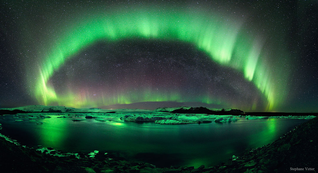 Ein grüner Polarlichtbogen wölbt sich im Bild, er spiegelt sich im Gewässer im Vordergrund.