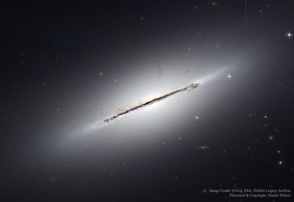 Die von der Seite sichtbare Galaxie sieht wie ein ovaler Nebel aus, der in der Mitte von einem dunklen, ausgefransten Staubband durchzogen ist.