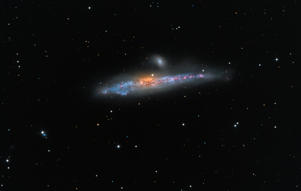 Im Bild schwebt eine leicht asymmetrische, von der Seite sichtbare Galaxie aus blauen Wolken, davor einige rosarote Staubwolken und in der Mitte eine orangefarbene Wölbung. Darüber leuchtet eine kleine ovale milchige Galaxie.