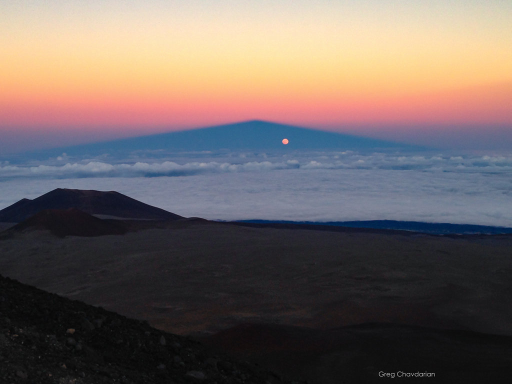 Hinter einem dunklen Gelände breitet sich eine Nebeldecke bis zum Horizont aus, dahinter steigt am Himmel im Abendrot der Schatten eines Vulkans auf. Im Schattenkegel leuchtet der aufgehende Mond.