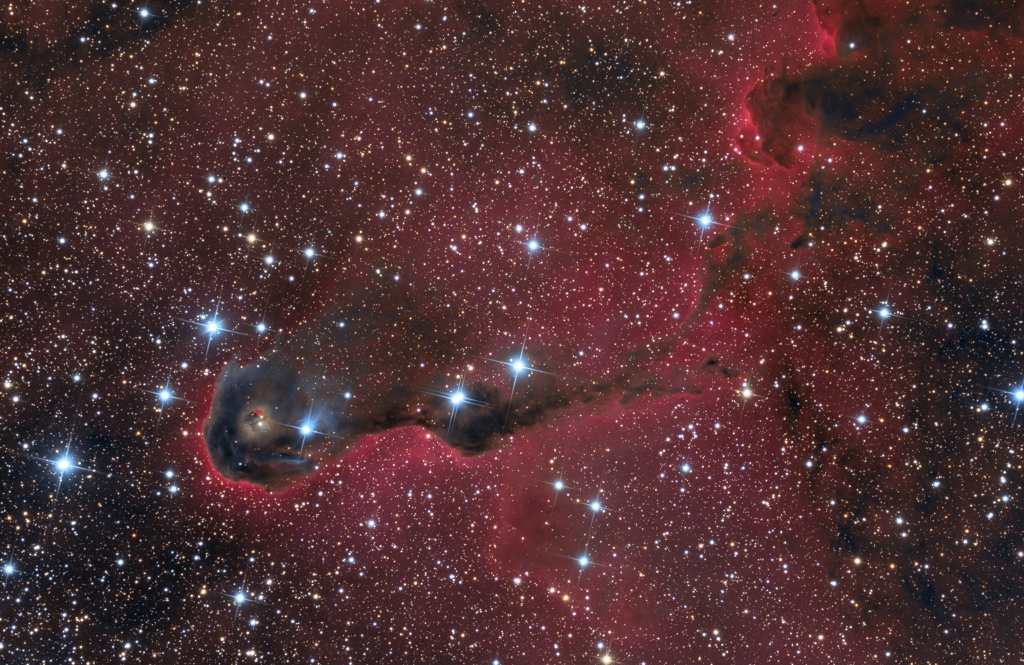 In der Mitte ist eine dunkle Wolke mit rot leuchtendem Rand und einigen hellen Sternen darin. Im Hintergrund sind viele Sterne auf schwach leuchtenden Nebeln verteilt.