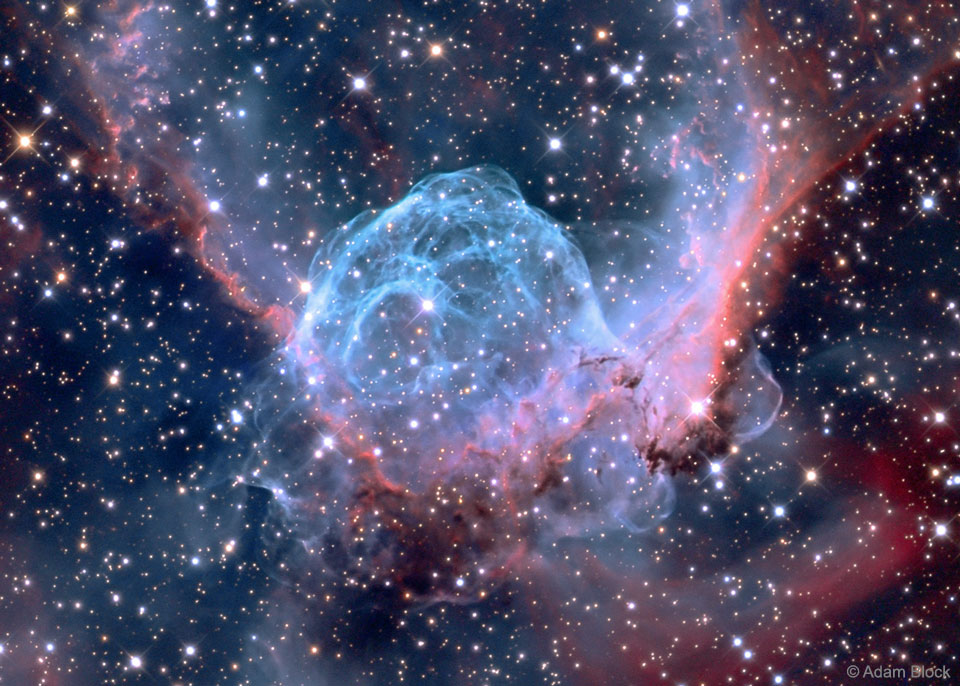 Der Nebel im sterngefüllten Bild hat viele schlierenartige Strukturen, er leuchtet blau und rötlich. In der Mitte ist eine Kuppe, links und rechts verlaufen Fortsätze nach oben, die an Flügel erinnern. Die Form erinnert an einen geflügelten Helm.