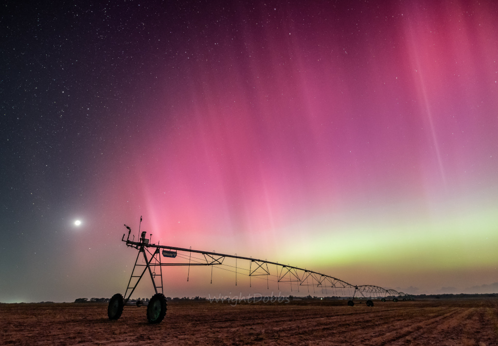 Über einem Feld mit einer Bewässerungsanlage schimmert am Himmel ein ausladendes Polarlicht. Unten verläuft ein grünlich-weißlicher Streifen, darüber ein breiter roter Bereich mit senkrecht gestreiften Polarlichtschleiern. Links leuchtet der Mond.