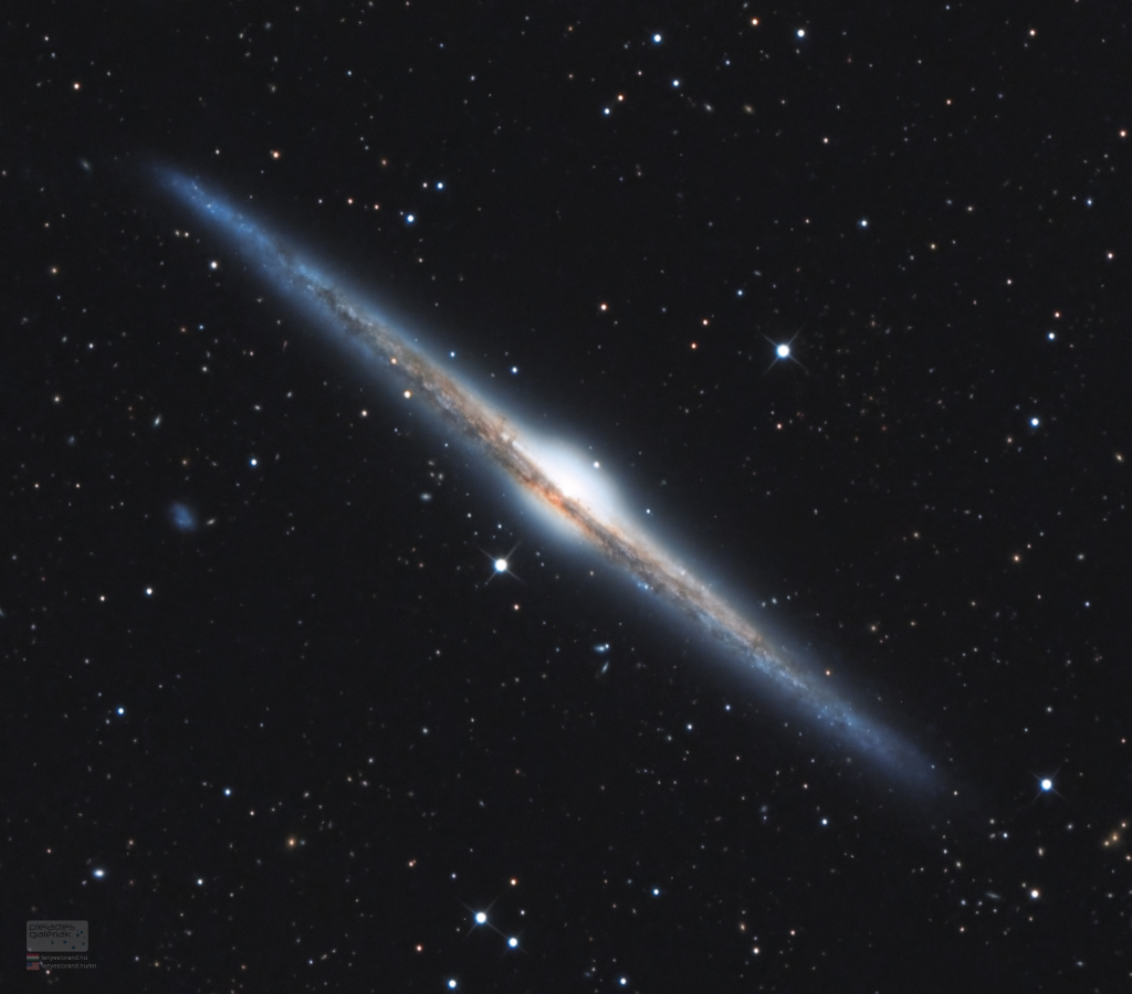 Die Galaxie im Bild ist von der Kante zu sehen und ganz schmal. In der Mitte ist eine helle Wölbung, außen verläuft ein Staubring die ganze Galaxie entlang.