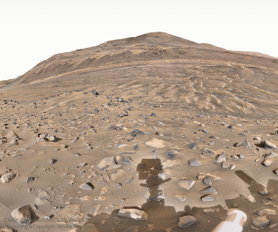 Der Schatten des Rovers Perseverance fällt auf eine steinige Marslandschaft. Der Rover befindet sich im Krater Jezero. Hinten am Horizont ragt ein Berg auf.