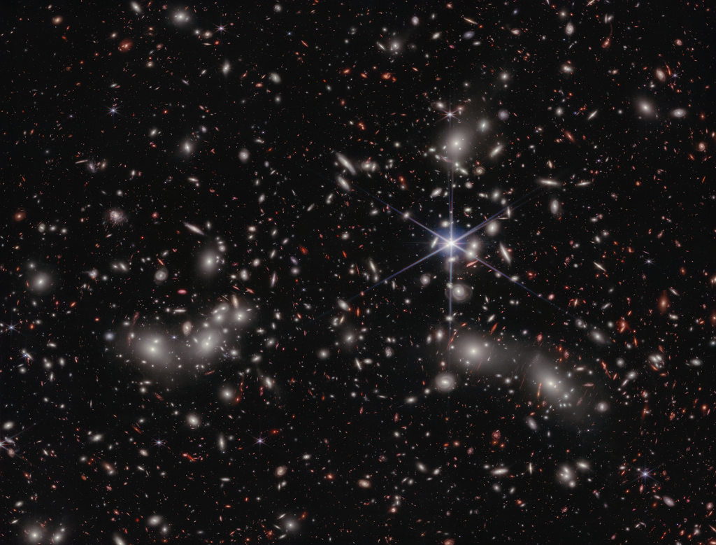 Das Bild ist voller Galaxien, es zeigt den Galaxienhaufen im Sternbild Bildhauer. Mitten im Bild ist ein Stern, der die Beugungesspitzen zeigt, die für das Weltraumteleskop Webb typisch sind.