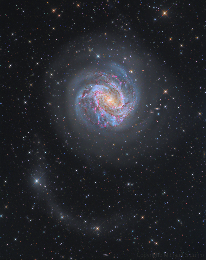 Die Galaxie in der Bildmitte ist direkt von oben zu sehen, sie hat einen gelblichen Kern, blaue Spiralarme und ist von Staubwolken und roten Sternbildungsregionen überzogen.