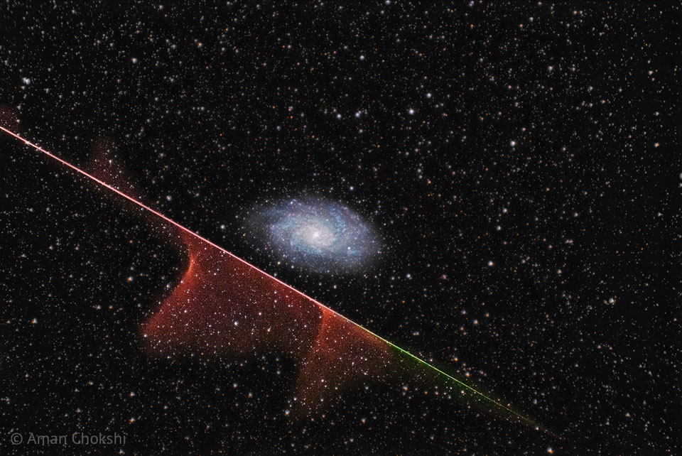 Mitten im Bild ist eine Spiralgalaxie, die Dreiecksgalaxie M33. Darunter zischt ein Meteor diagonal durchs Bild, links oben leuchtet die Spur rot, rechts unten grün, nach unten strömen nebelartige Schwaden von der Spur aus.