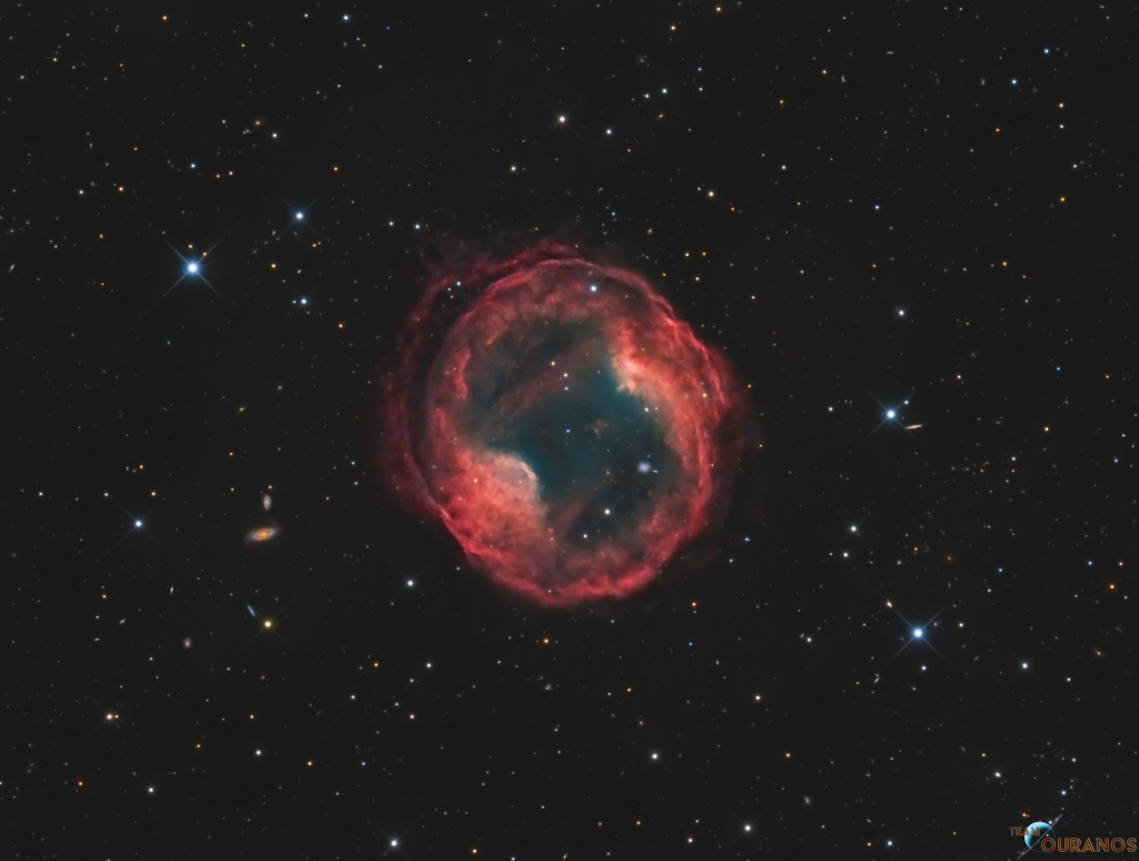 Der planetarische Nebel Jones-Emberson 1 ist eine rot leuchtende Hülle mit einem dunklein Inneren. Der Nebel schwebt mitten im Bild und ist von dünn verteilten Sternen und Galaxien umgeben.