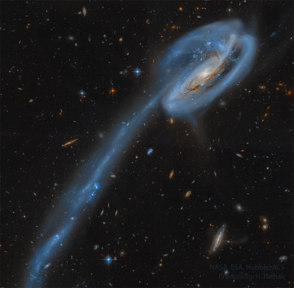 Rechts oben ist eine Spiralgalaxie, die von schleifenförmigen Spiralarmen umwickelt ist. Nach links unten zieht sich ein langer Gezeitenschweif aus blauen Sternhaufen.