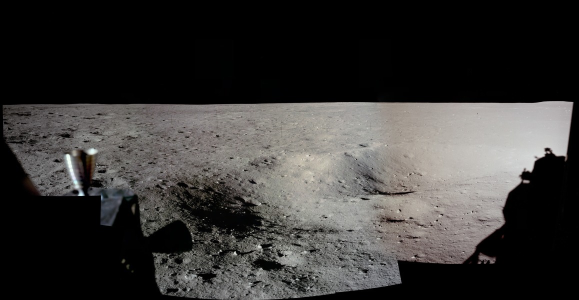 Der Blick fällt auf eine graubraune Einöde mit kleinen Kratern und Gesteinsbrocken. Links sind Schubdüsen der Landefähre Eagle, rechts fällt der Schatten des Adlers auf den Mondboden.