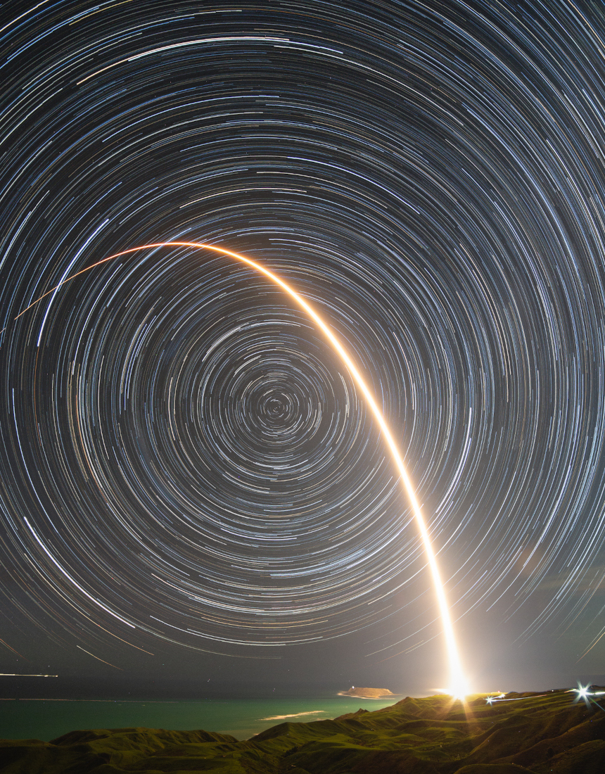 Vor einem Himmel voller Strichspuren von Sternen leuchtet der Lichtbogen einer startenden Rakete. Unten ist das grünliche Wasser an der Küste zu sehen.