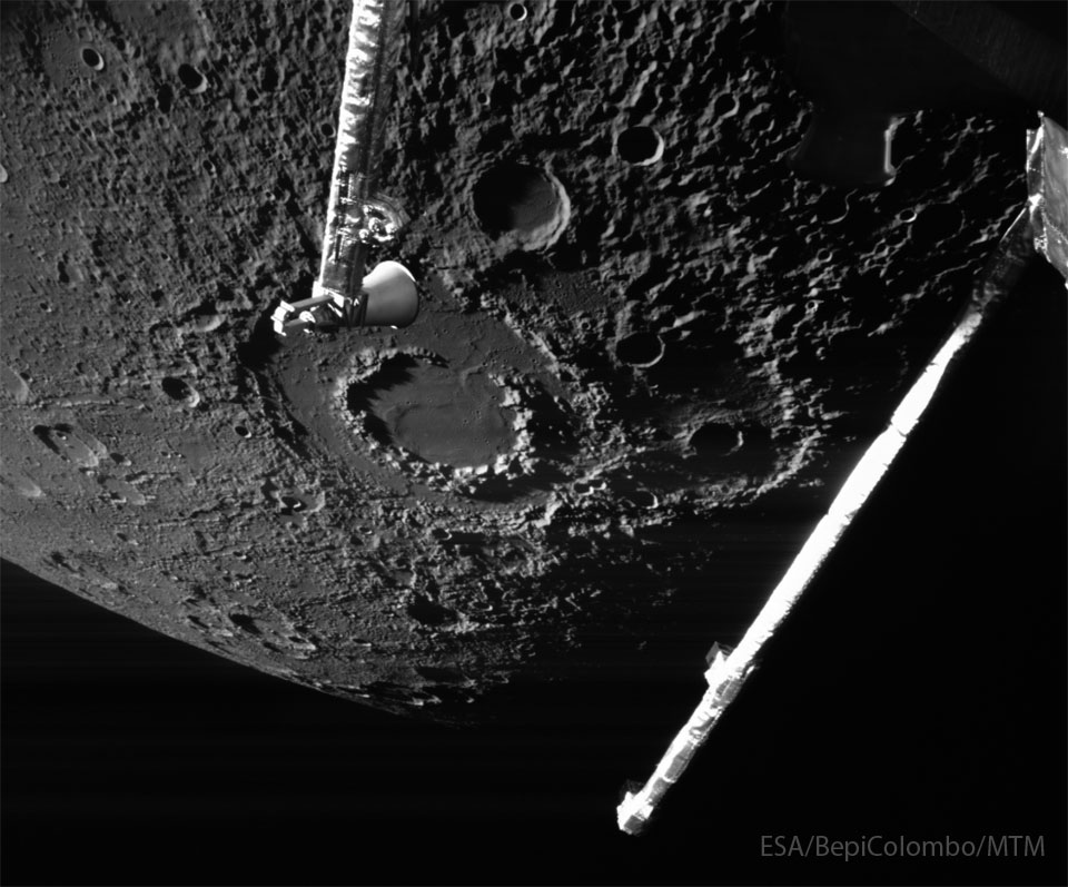 Hinter zwei Auslegern der Raumsonde BepiColombo ist die Oberfläche des Planeten Merkur zu sehen. Der Rand des Planeten ist unten. Die Oberfläche ist von Kratern übersät, in der Bildmitte liegt der markante Krater Vivaldi mit seinen zwei konzentrischen Kraterwällen. 