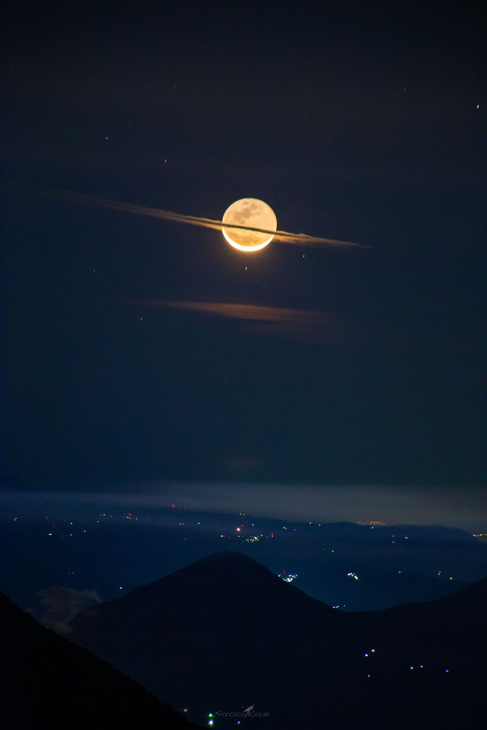 Über einem dunklen Vulkangipfel leuchtet der Mond, die Sichel unten ist direkt im Sonnenlicht, die Nachtseite wird von der Erde beleuchtet. Vor dem Mond ist eine Wolke, die an Saturnringe erinnert.