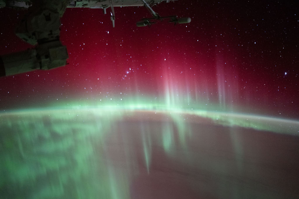 Von der Erde steigen grüne Strahlen auf, darüber ist ein breites diffuses rotes Schimmern. Durch das Polarlicht hindurch sind Sterne zu sehen. Links oben ragt ein Teil der Internationalen Raumstation ins Bild.