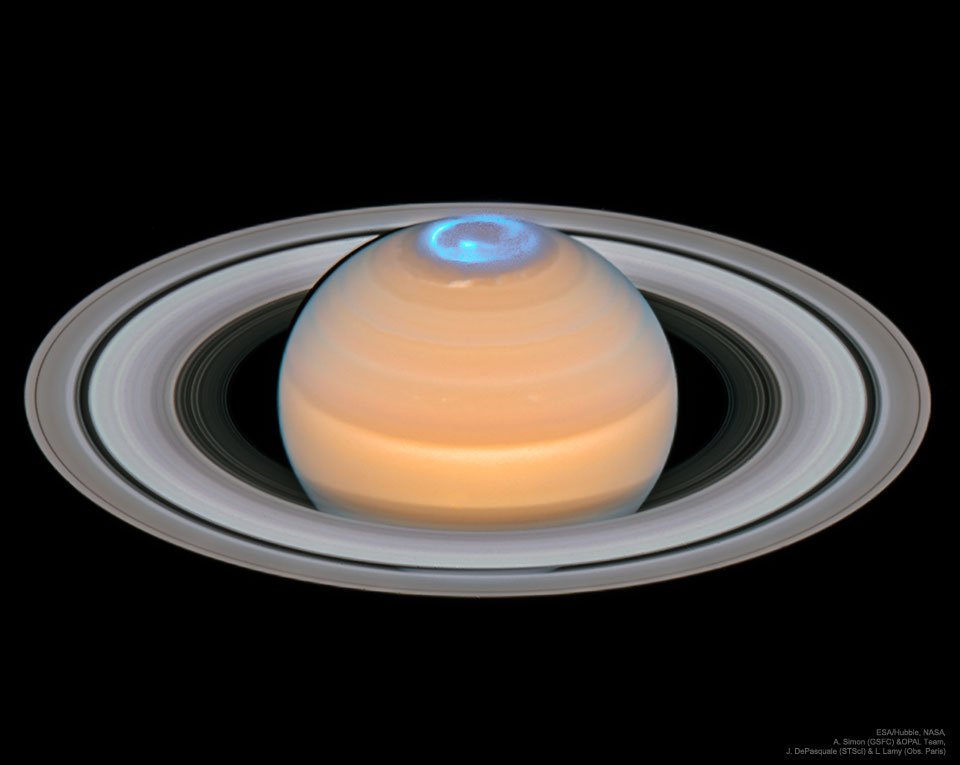 Wir sehen Saturn schräg von oben, die Ringe breiten sich übers ganze Bild aus, sie sind oben und unten breiter als der Planet. Am Pol des Planeten leuchten cyanfarbene Polarlichter in Form einer Spirale.