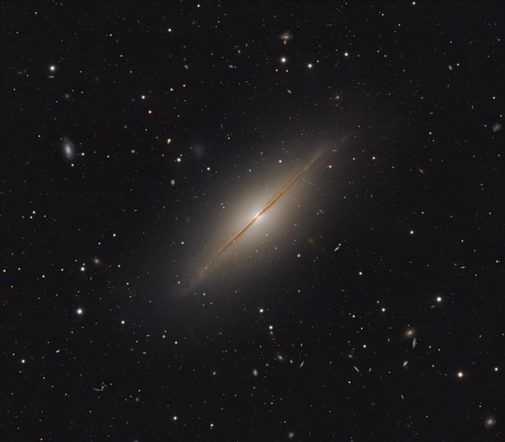 Mitten im Bild ist eine nebelartige Galaxie, die wir von der Kante sehen. Sie liegt schräg im Bild, umgeben von vielen weiteren Galaxien, die wie Sterne im Bild verteilt sind. Ein Staubwulst schneidet scheinbar die Galaxie in der Mitte durch.