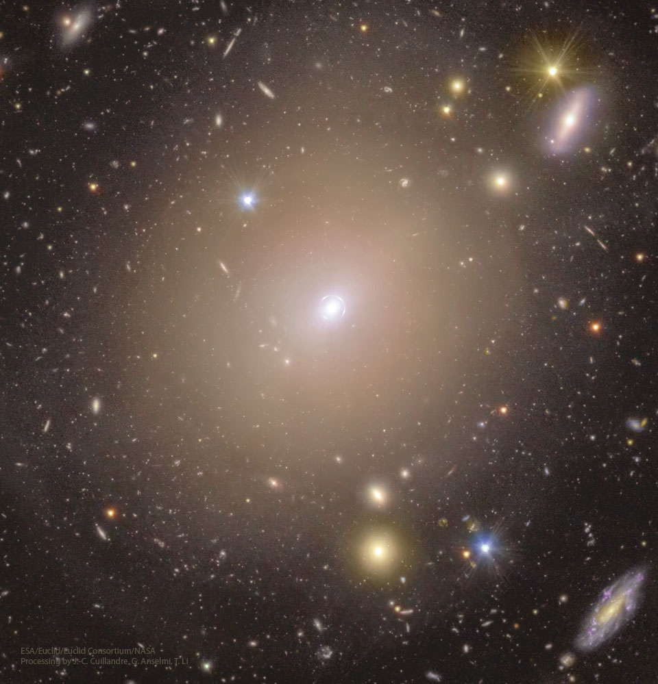 Fast das gesamte Bild ist von der durscheinenden gelblichen Sternenwolke einer elliptischen Galaxie erfüllt. Um ihr weißlich strahlendes Zentrum verläuft ein ebenso strahlender, kleiner Kreis. Im Hintergrund stehen unzählige weitere, kleine Galaxien in verschiedenen Formen und Farben. Ein Klick auf das Bild lädt die höchstaufgelöste verfügbare Version.