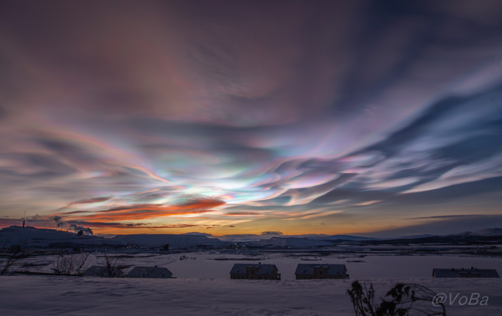 Über einer verschneiten Landschaft mit einigen Häusern im Vordergrund wabern bunt schillernde Wolken.