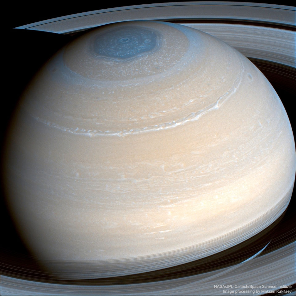 Die Saturnkugel ist von beigefarbenen Wolkenringen gemustert. Oben ist ein blassblaues sechseckiges Wolkenmuster. Oben ist ein Teil der Ringe zu sehen, links oben fällt der Schatten des Planeten Saturn auf die Ringe.