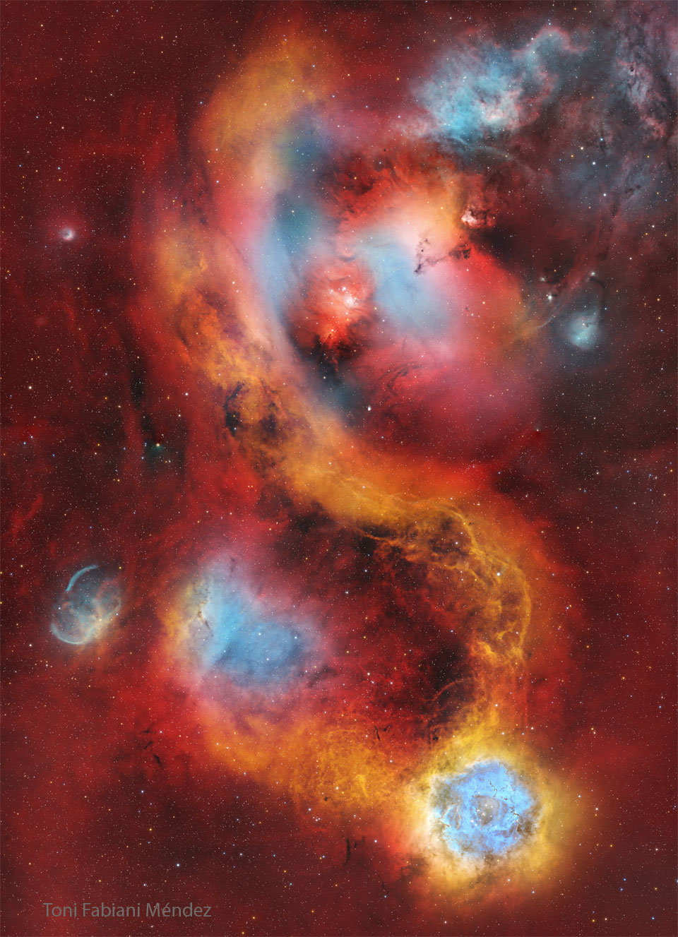 Das Bildfeld ist voller rot leuchtender Nebelschwaden. Dazwischen sind orangefarbene und blaue Wolken verteilt. Rechts unten ist der berühmte Rosettennebel.