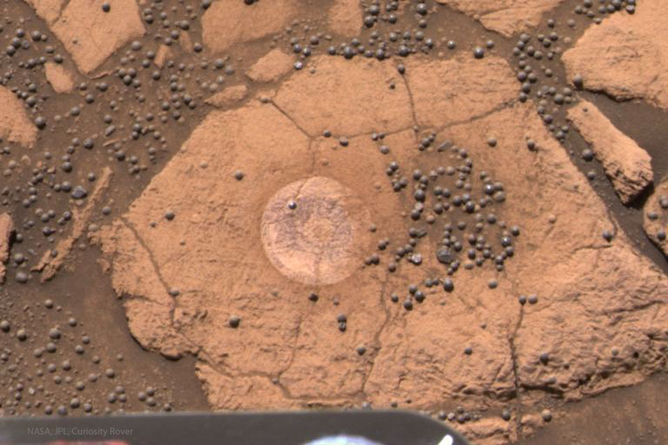 Mitten im Bild ist ein großer, rotbrauner Stein. Rechts liegen viele "Blaubeeren" in einer Senke, links daneben nahm der Rover Opportunity eine runde Probe. Außen um den Stein ist dunkles Fugenmaterial zwischen weiteren hellen Steinen verteilt, auf dem viele Blaubeeren liegen.