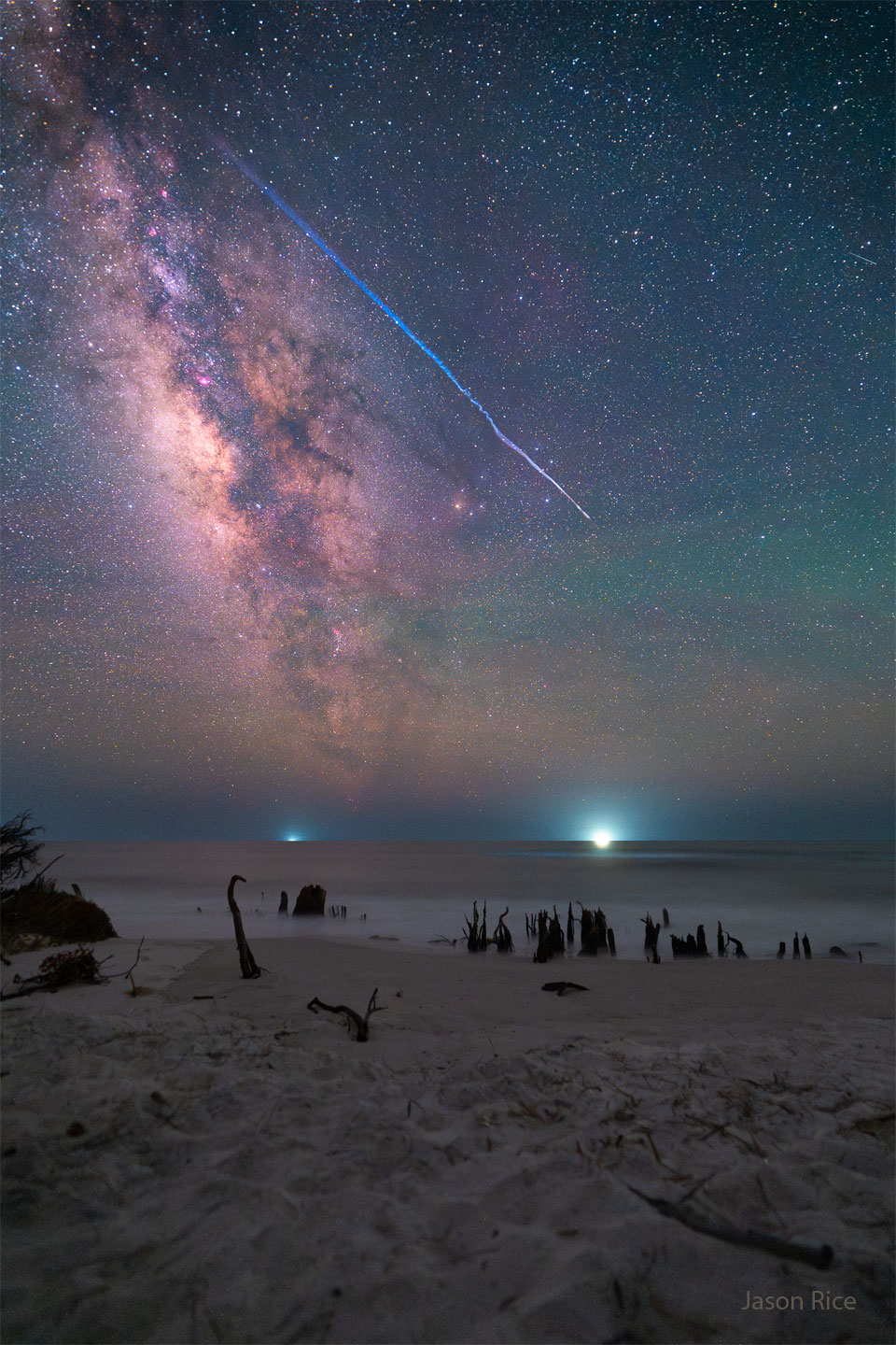 Über den Sternenhimmel an einem Strand mit toten Baumstämmen zischt ein heller Streifen. Wenn man mit der Maus über das Bild fährt, kommt die Rauchspur zutage, die der helle Meteor hinterlassen hat.