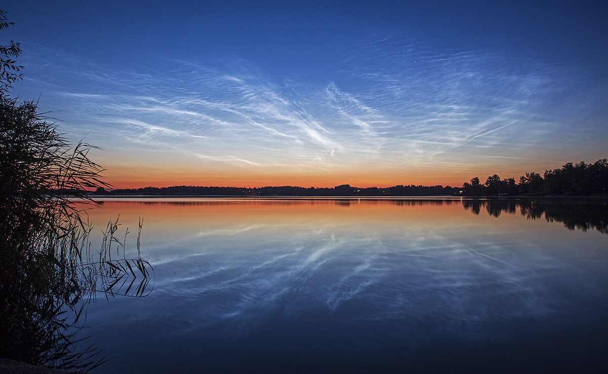 Über einem See in Schweden leuchten Wolken. Sie sind am Rand des Weltraums, daher leuchtet die Sonne noch darauf, obwohl es am Boden schon dunkel ist. Unten ist ein See, der die Wolken spiegelt.