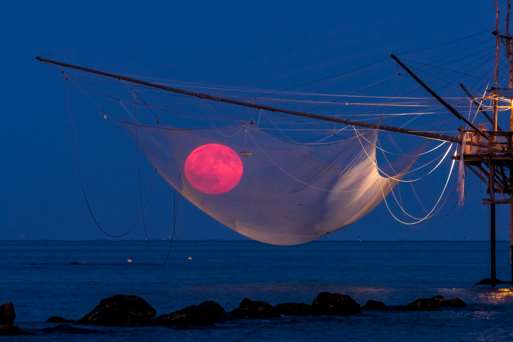 In den Netzen eines alten Fischerbootes hängt der rote Mond am blauen Himmel. Dahinter reicht das Meer bis zum Horizont.