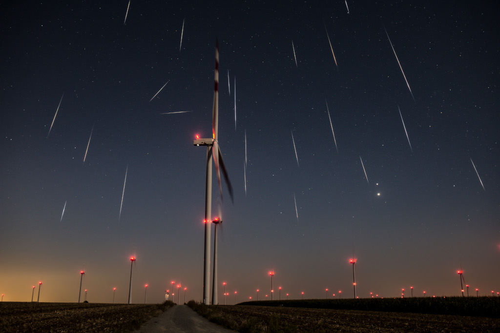 Über einem Windpark im Burgenland, dessen Lichter rot blinken, regnen Meteore der Perseïden vom Himmel.