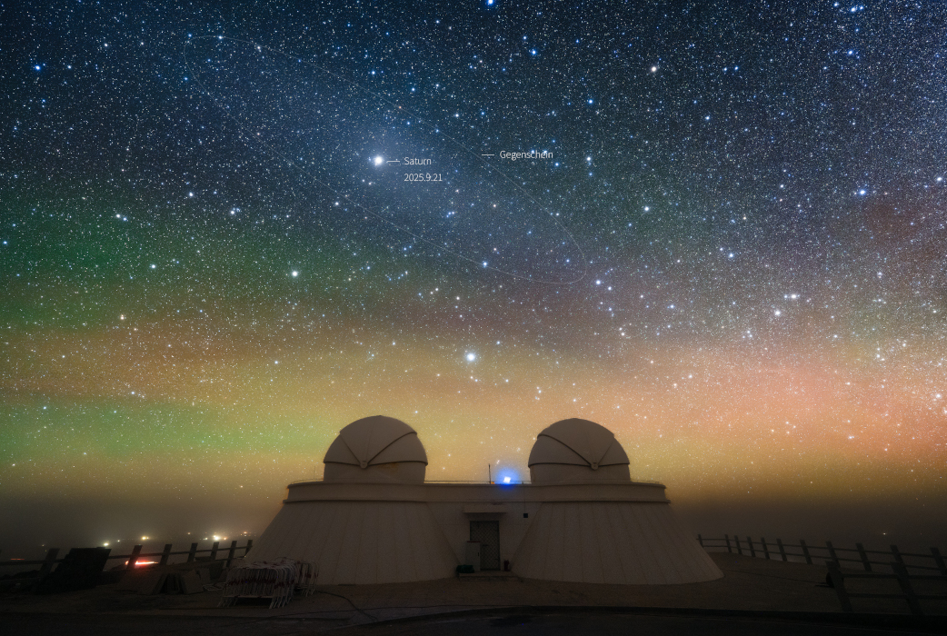 Über den Kuppeln eines Zwillingsteleskops breitet sich ein dichter Teppich aus Sternen aus. Unten ist der Himmel hell und rosa bis grünlich gefärbt. Oben steht der Planet Saturn. Er ist vom Gegenschein umgeben.