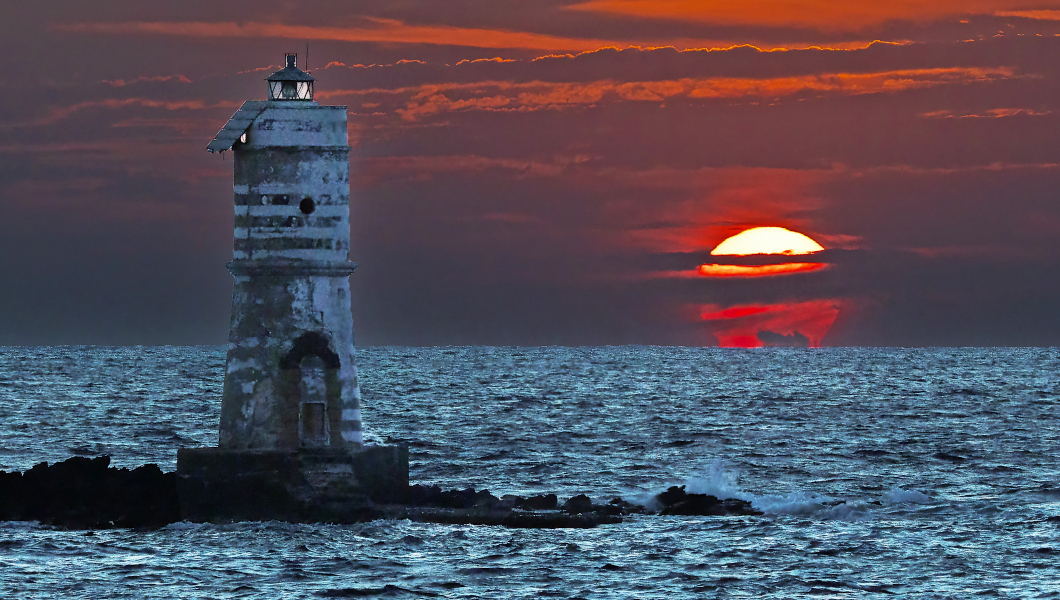 Hinter dem Leuchtturm von Mangiabarche geht die Sonne unter. Der Blick reicht über das Meer bis zum Horizont. Die Sonne ist von dunklen Wolken gestreift.