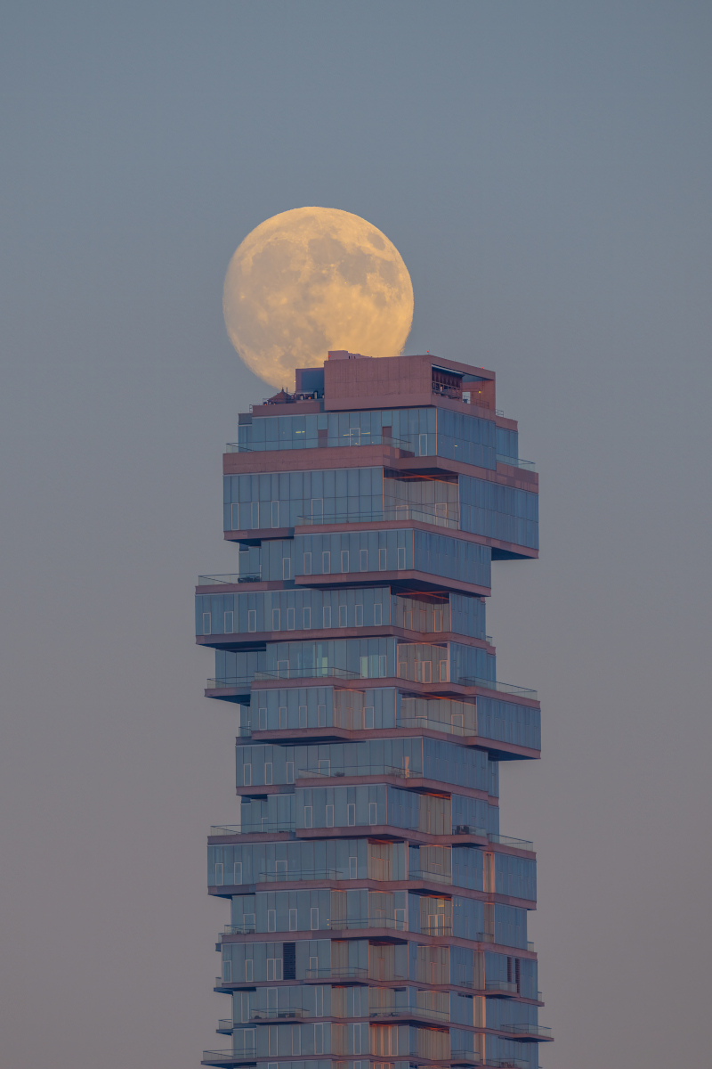 Der Vollmond geht über einem Gebäude auf, das an einen Jenga-Turm erinnert. Er leuchtet gelb am noch hellen Himmel in der Dämmerung, der vom Erdschatten grau gefärbt ist.