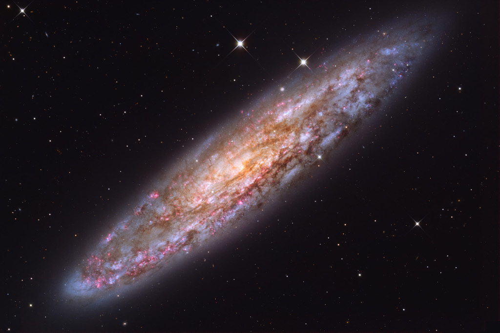 Die Galaxie NGC 253 liegt schräg im Bild. Ihre Oberfläche ist von dunklen Staubwolken, blauen Sternhaufen und roten Sternbildungsregionen überzogen. Ihre Form erinnert an eine Münze.