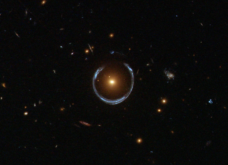 Um eine elliptische gelbe Galaxie biegt sich ein blauer Ring. Es ist das verzerrte Abbild einer Galaxie, die viel weiter entfernt ist. Die gelbe Galaxie im Vordergrund wirkt dabei wie eine Linse, die das Licht bricht.