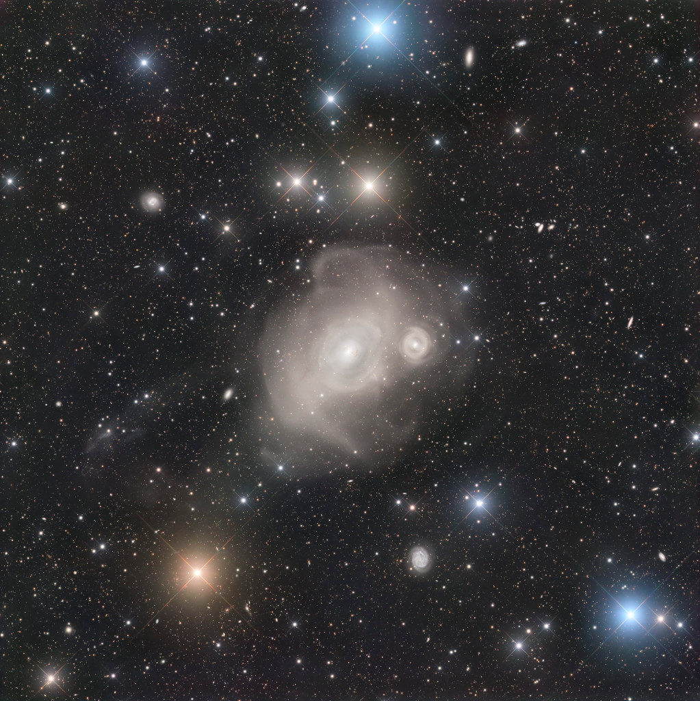 Eine große Galaxie ist von schalenartigen Hüllen umgeben. Rechts neben ihrem Zentrum ist eine kleinere Galaxie mit Hüllen. Beide gehören zum Fornax-Galaxienhaufen. Im Bild sind einige weitere, kleinere Galaxien und ein paar Sterne mit Lichtkreuzen verteilt. Dazwischen sind viele kleine Sterne.