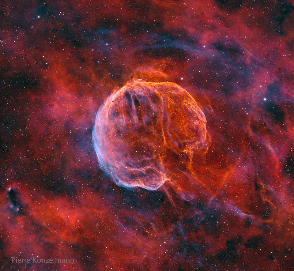 Ein kompakter roter Nebel leuchtet mitten im Bild. Er ist von roten Nebelschwaden und einem zarten, fast transparenten blauen Schleier umgeben. Seine Form und Struktur erinnern an ein Gehirn.
