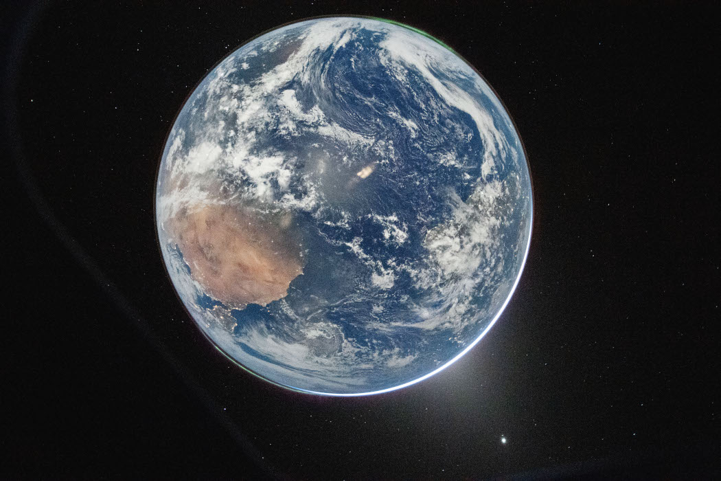 Vor einem schwarzen Hintergrund mit wenigen Sternen steht die Erde als blaue Scheibe mit wenigen weißen Wolken. Links ist eine rotbraune Landmasse zu erkennen.