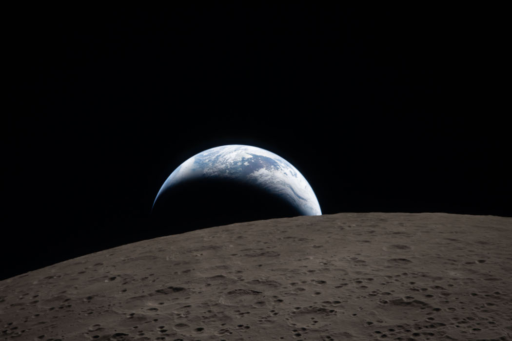Über dem Horizont des dunklen Mondes, der von Kratern bedeckt ist, steht die Erde als breite Sichel. Man erkennt weiße Wolken und blaue Meere.