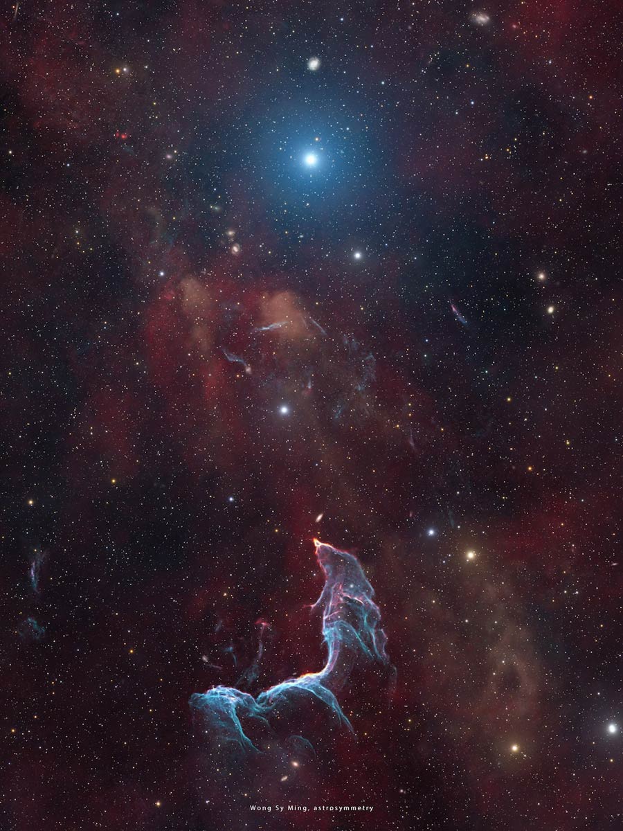 Unter einem hellen, blau strahlenden Stern windet sich eine zart umrissene Gestalt. Manche erkennen darin eine Meerjungfrau, andere einen Kampffisch. Im Bild sind auch einige kleine Galaxien erkennbar. Der Hintergrund ist von winzigen Sternen übersät, dazwischen leuchten einige hellere Sterne.